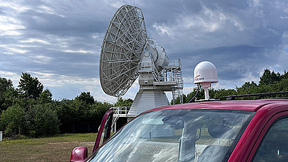 Satellite Dish and Vehicle with Antenna A large satellite dish in an open field with a red vehicle in the foreground. The vehicle is equipped with a small white dome-shaped antenna. Dark clouds are visible in the sky, suggesting incoming weather changes.
